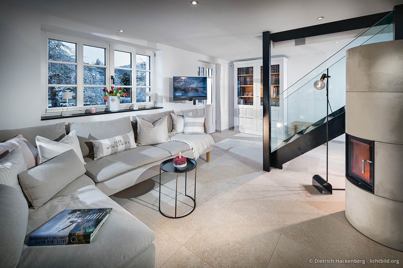 Sofaecke mit Fensterblick auf Schneelandschaft, Speicherofen und Treppenaufgang mit gläsernem Geländer. Sauerland Ferienhaus - Fachwerkhaus Eiben, Benolpe