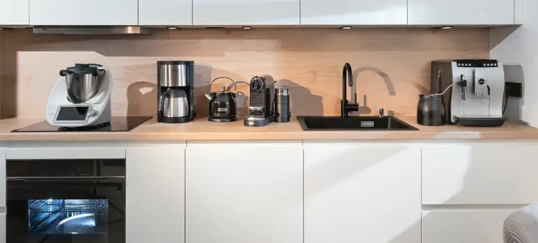 Drei Varianten von automatischen Kaffeebereitern (Kaffeevollautomat, Nespresso- & Filterkaffeemaschine), Wasserkocher und Thermomix auf der Arbeitsfläche der modernen Einbauküche.