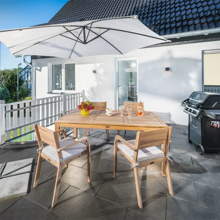 Terrasse in der Morgensonne mit Holzesstisch mit Stühlen, Sonnenschirm und Grill